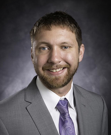 Portrait of Charles Webb - a man with brown hair wearing a gray suit and purple tie, smiling at the camera