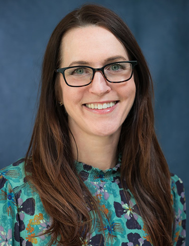 Portrait of Jennifer Schaff - a woman with brown hair, glasses, and a multicolored shirt smiles at the camera