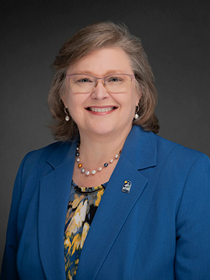 Woman wearing glasses, a blue blazer, and a patterned blouse, photographed against a blue gradient background.