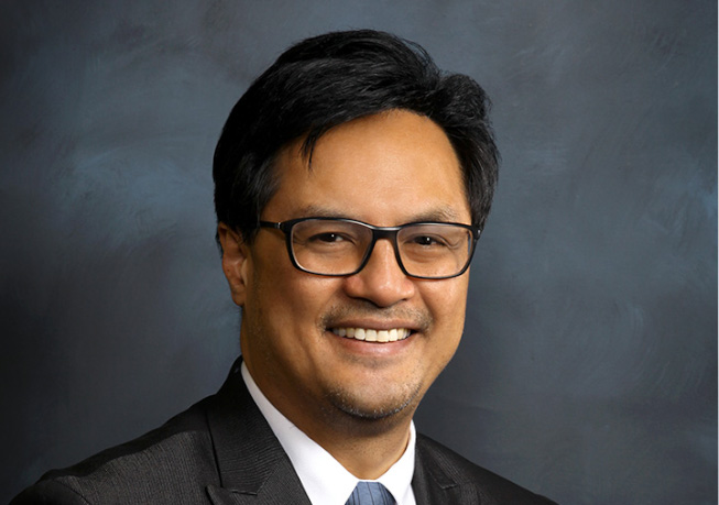 Studio headshot of an adult professional wearing glasses, a dark suit jacket, and a light-colored shirt, set against a dark textured background.
