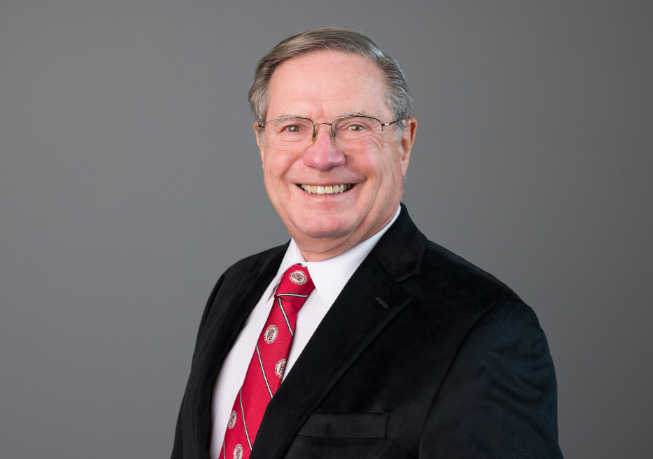 Studio headshot of an adult professional wearing a dark suit jacket, white shirt, and red patterned tie, photographed against a medium gray background.