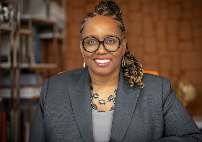 Professional portrait of an adult wearing a dark blazer and statement necklace, seated indoors with shelving and warm-toned decor in the background.