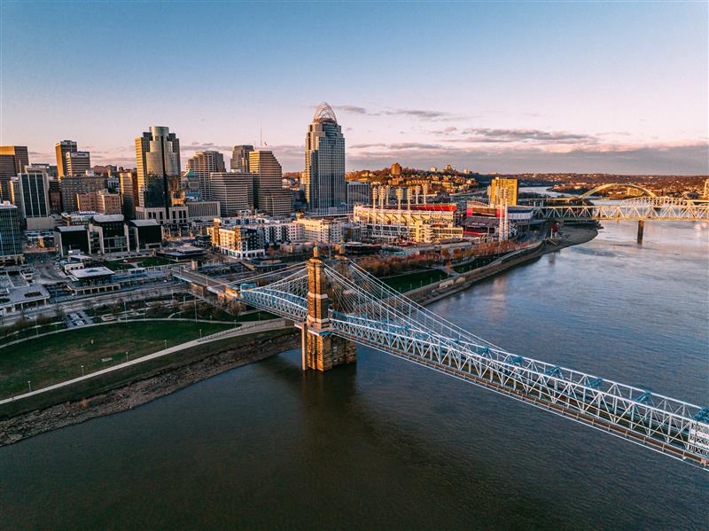 Skyline along the Ohio River featuring the John A. Roebling Suspension Bridge in the foreground and a riverfront stadium and downtown buildings beyond