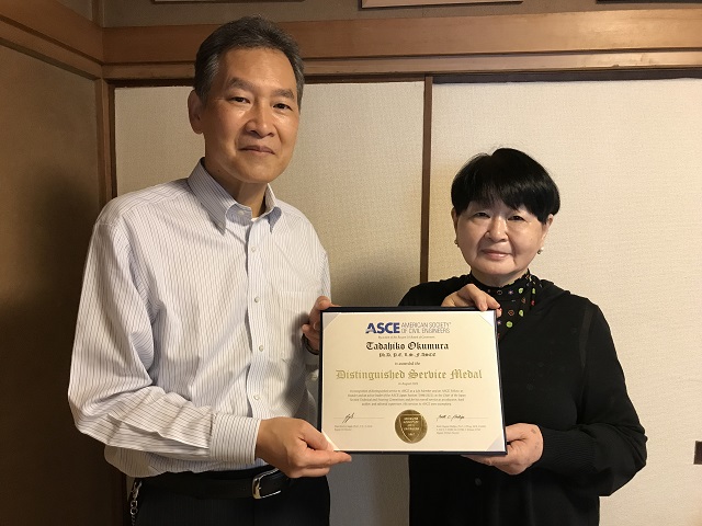 The family of the late Dr. Okumura accepted the award on his behalf. Pictured are two people holding the award and looking at the camera smiling. 
