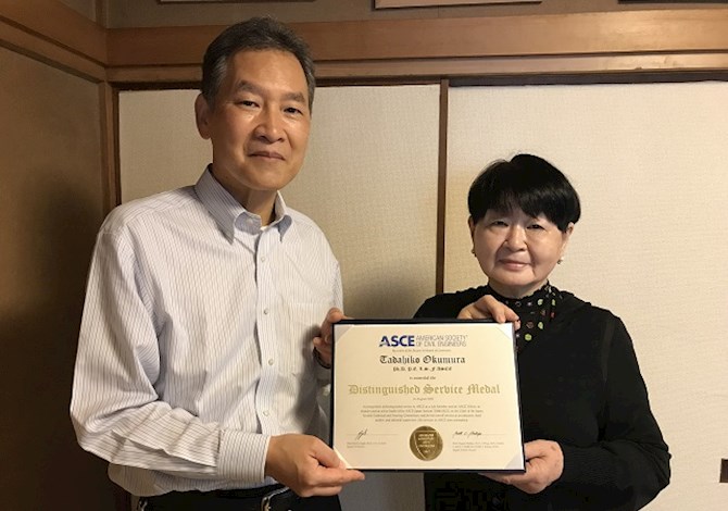 The family of the late Dr. Okumura accepted the award on his behalf. Pictured are two people holding the award and looking at the camera smiling.
