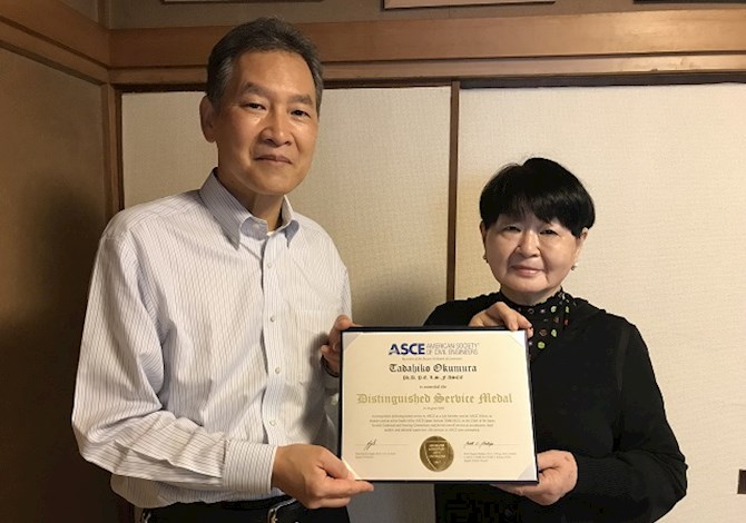 The family of the late Dr. Okumura accepted the award on his behalf. Pictured are two people holding the award and looking at the camera smiling. 