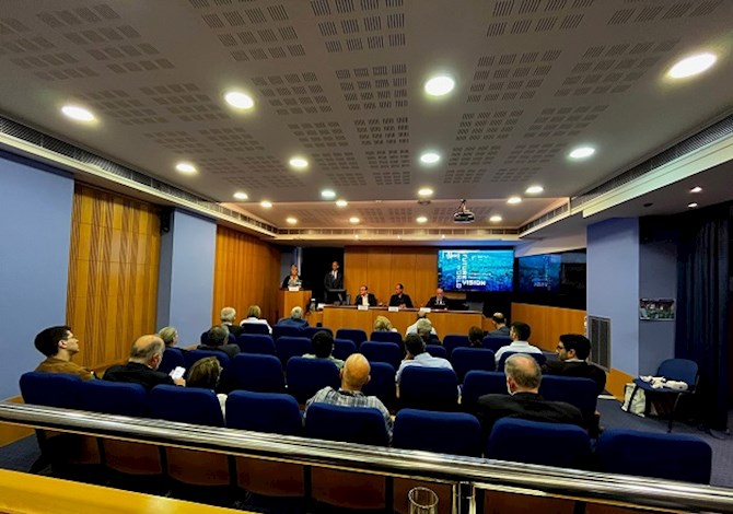 Pictured: Eighteen people sitting in an auditorium watching the presentation given by five presenters in the front of the room next to a screen. 