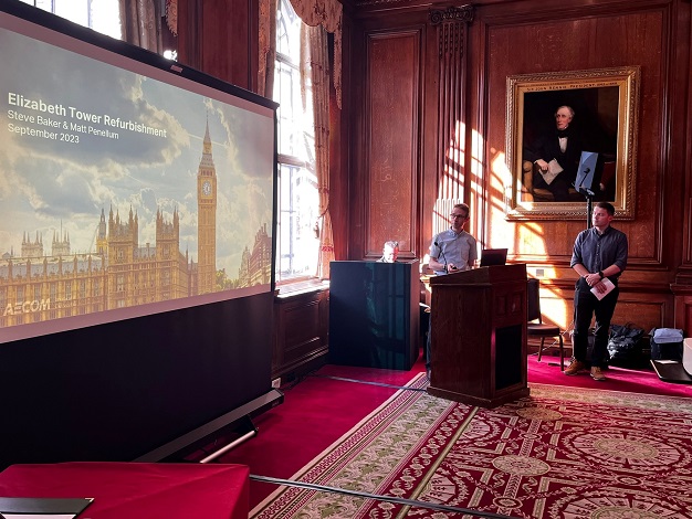 Pictured: One person standing at a podium and one person standing to the right of the podium giving a presentation next to a screen. 