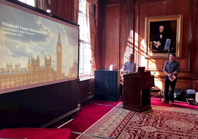 Pictured: One person standing at a podium and one person standing to the right of the podium giving a presentation next to a screen. 