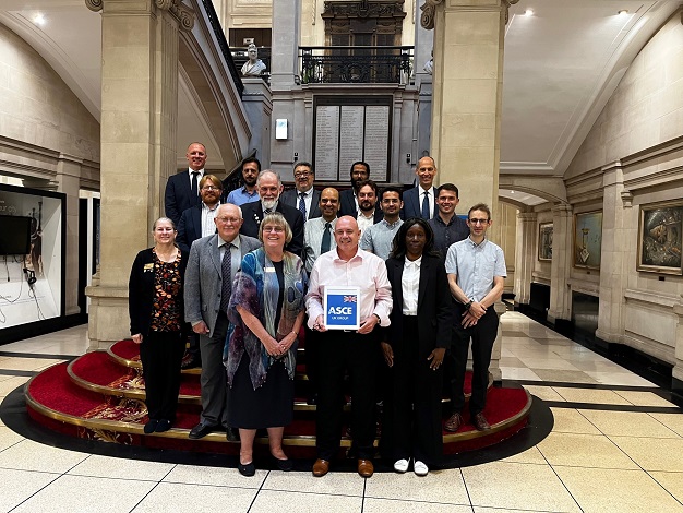Pictured: Seventeen people standing on stairs in front of a plaque smiling at the camera