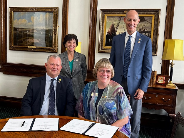 Pictured: Two people sitting at a desk and two people standing behind them smiling at the camera. 