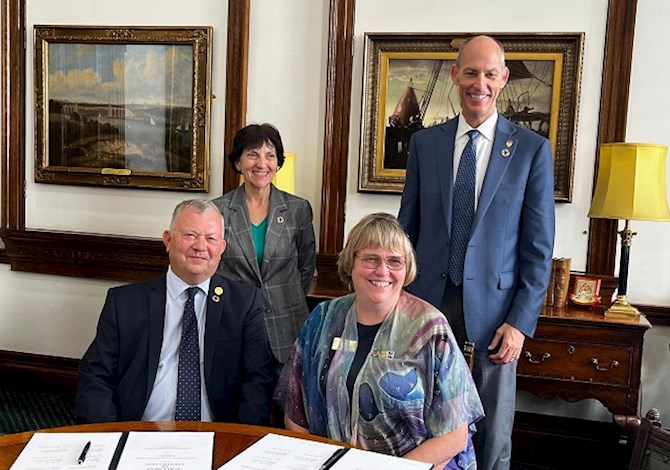 Pictured: Two people sitting at a desk and two people standing behind them smiling at the camera.