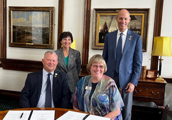 Pictured: Two people sitting at a desk and two people standing behind them smiling at the camera. 