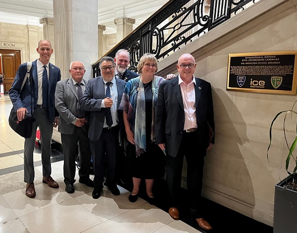 Pictured: Six people smiling at the camera in front of the stairs and next to a plaque. 