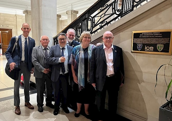 Pictured: Six people smiling at the camera in front of the stairs and next to a plaque.
