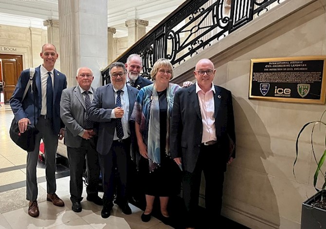 Pictured: Six people smiling at the camera in front of the stairs and next to a plaque. 