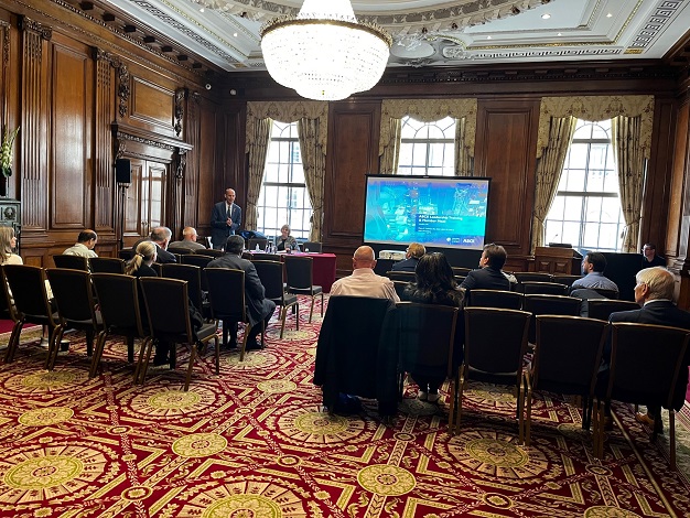 Pictured: fourteen people sitting in a conference room, looking at a presentation on a screen with two people in the front of the room presenting. 
