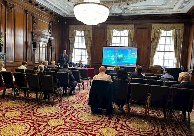 Pictured: fourteen people sitting in a conference room, looking at a presentation on a screen with two people in the front of the room presenting. 