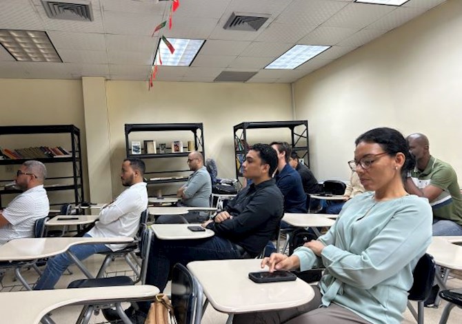 Eight people are sitting at desks watching a presentation