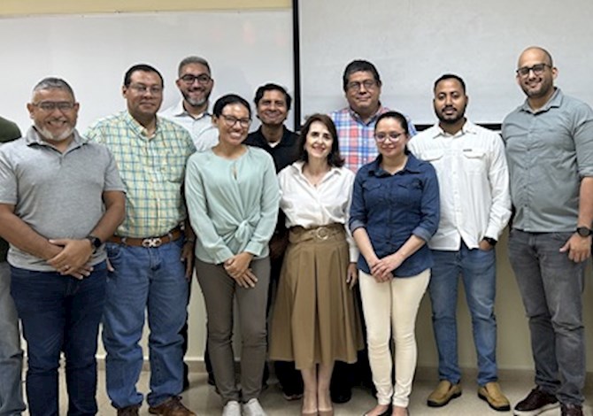 Fourteen people are standing in front of a screen smiling at the camera