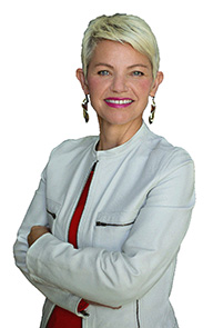 Woman with short blond hair wearing a white leather jacket and pink shirt smiles at the camera.