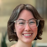 Woman with brown hair wearing a greenish brown blouse and glasses smiles at the camera. 