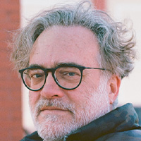 Man with graying hair and beard wearing glasses and a puffer jacket looks at the camera. 