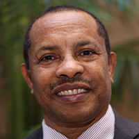 Man with brown skin in jacket and tie smiles at the camera. He has a mustache.