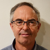 A man wearing glasses and a denim shirt smiles at the camera. 