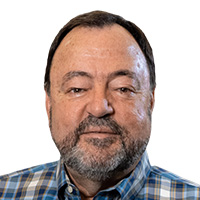 A man with a greying beard, wearing a blue plaid shirt, looks out at the camera. 