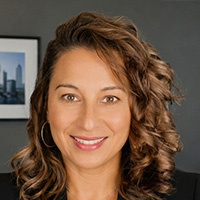 A woman with brown curly hair and wearing hoop earrings smiles at the camera. 