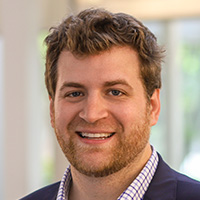 A man wearing a plaid shirt and a suit jacket smiles at the camera. 