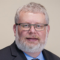 Man with cropped hair and bead is wearing glasses and smiling at the camera. He has on a blue suit and a patterned tie.