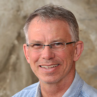 A man wearing glasses and a light-colored denim button-down shirt smiles at the camera. 