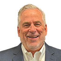 Man with graying hair and beard smiles at the camera. He’s wearing a button-up shirt and a gray suit. 