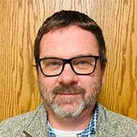 A man wearing glasses, a plaid shirt, and a fleece vest smiles at the camera. 