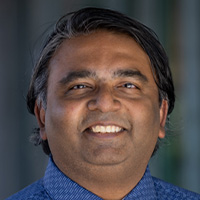 A man with brown skin and black hair wearing a blue button-down shirt smiles at the camera. 