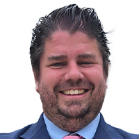 A man in a blue suit, blue shirt, and pink tie, his hands clasped before him, smiles at the camera. 