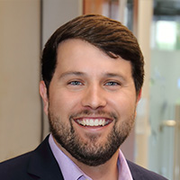 Man with short brown hair wearing a lavender shirt and black jacket smiles at the camera. 