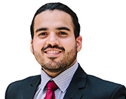 Man with dark brown hair wearing a dark blue suit with a blue patterned shirt and maroon tie.