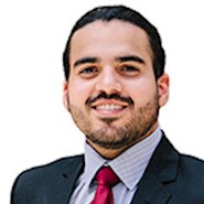 Man with dark brown hair wearing a dark blue suit with a blue patterned shirt and maroon tie.