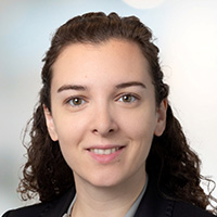 Woman with dark brown curly hair has on a gray blouse and black jacket. 