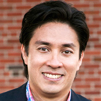 Man in plaid shirt and blazer smiles at the camera.