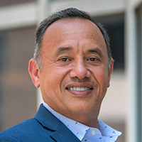 A man in a blue suit with a blue shirt and blue tie smiles at the camera. 