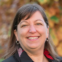 A woman with blondish-brown hair is wearing a floral blouse underneath a black blazer. 
