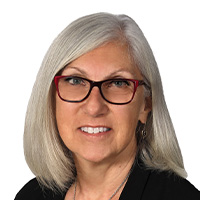 Woman with shoulder-length blonde hair wearing a black jacket over a black and white blouse smiles at the camera. 