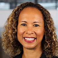Woman with brown skin and curly brown and blonde hair smiles at the camera. 