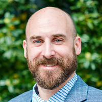 Man in blue suit jacket and blue and white striped shirt smiles at the camera. 