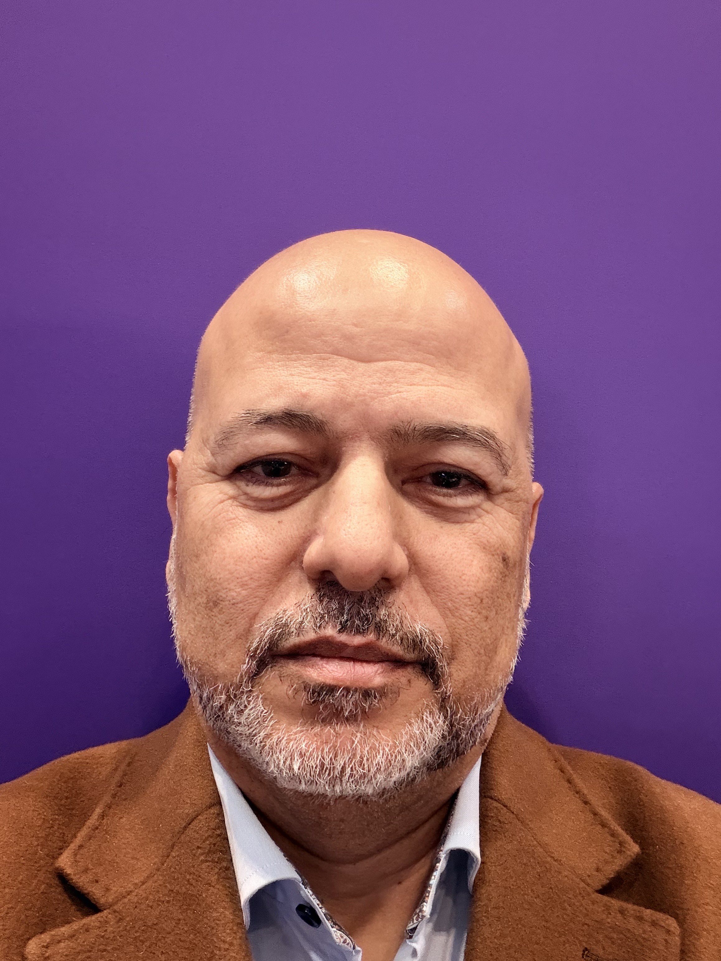 Man with brown skin and a goatee smiles at the camera. He is wearing a brown coat with a blue shirt. 
