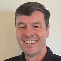 A man wears a black polo shirt with writing on it smiles at the camera. 